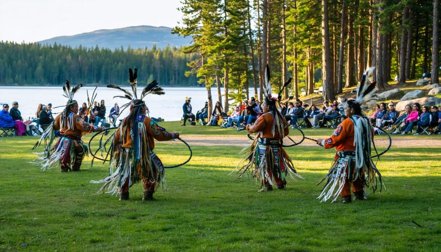 Indigenous dancers in colorful regalia performing in a grassy park dance circle at golden hour, with spectators seated on blankets and chairs and white pines and a lakeshore in the background.