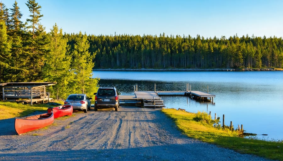 Gravel parking area with a few cars beside a calm lake, canoe rack and wooden dock, surrounded by spruce and birch forest at sunrise, quiet and uncrowded scene.