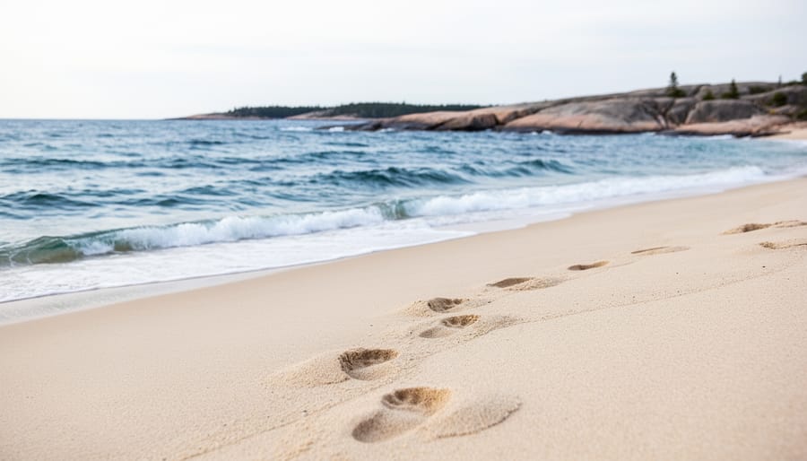 Wide photo of a sandy Georgian Bay shoreline with footprints, gentle waves, and a distant rocky coast under an overcast sky.